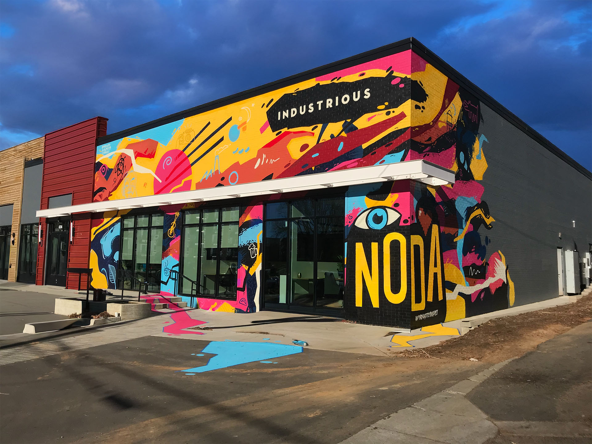 A colorful mural wraps around a two story building. The words Industrious are written on the top and NODA is written on the corner. 